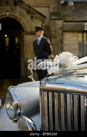 Nahaufnahme eines Jaguar Autos, das für eine Hochzeit mit weißen Bändern geschmückt ist, mit einem außer Fokus geratenden Chauffeur, der vor einer Kirche in der Ferne steht. Stockfoto