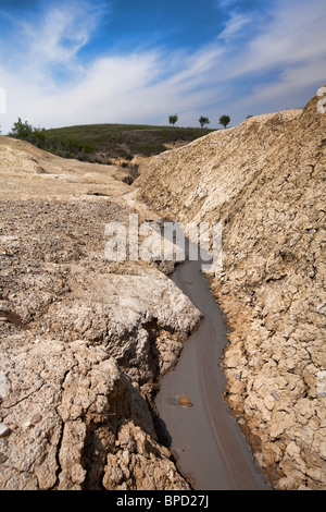Ein schmaler, sich windender Bach aus matschigem Wasser fließt durch ein tiefes, gerissenes, trockenes Flussbett, das in hellbraune Erde gehauen ist. Die Ufer sind steil und erodiert und weisen Schichten aus verdichtetem Boden und Gestein auf. Im Stockfoto