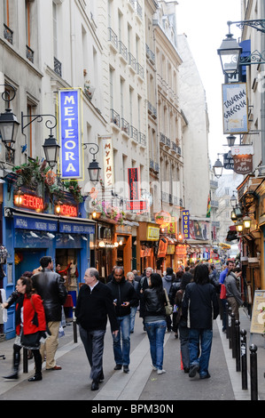 Rue Mouffetard Market Street Fußgänger beleuchtete Geschäfte Paris Frankreich | PARIS, Frankreich – Fußgänger spazieren vorbei an beleuchteten Restaurantschildern und griechischen und französischen Restaurants entlang der Rue Mouffetard, einer der ältesten Marktstraßen der Stadt. Durch das Quartier Latin im 5. Arrondissement verläuft die enge, sanft geschwungene Straße, die seit römischer Zeit genutzt wird und nach Einbruch der Dunkelheit einen lebendigen, traditionellen Charakter bewahrt. Neonschilder und Laternen-ähnliche Straßenlaternen beleuchten die Fassaden in der Abenddämmerung. Stockfoto