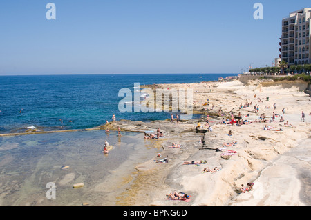 Sonnenanbeter entspannen Sie sich auf den steinigen Strand in Sliema, Malta. Links befindet sich ein Pool mit Meerwasser, der einen sicheren Bereich zum Plantschen bietet Stockfoto