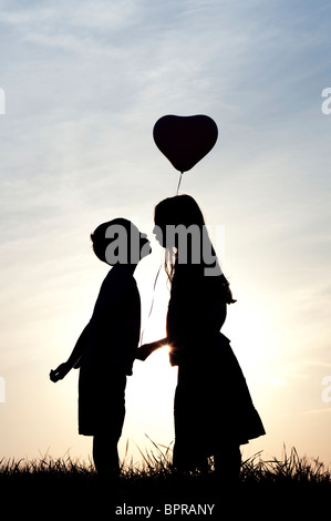 Silhouette der ein junges Mädchen und jungen Händchenhalten und küssen mit einem Herz-Form-Ballon bei Sonnenuntergang Stockfoto