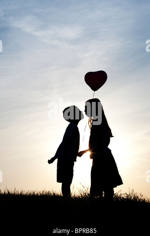 Silhouette der ein junges Mädchen und jungen Händchenhalten und küssen mit einem Herz-Form-Ballon bei Sonnenuntergang Stockfoto