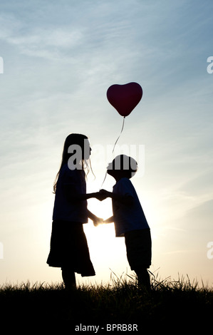 Silhouette der ein junges Mädchen und jungen Händchenhalten und küssen mit einem Herz-Form-Ballon bei Sonnenuntergang Stockfoto