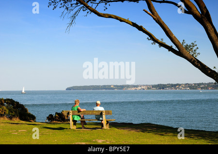 Zwei Personen sitzen auf der Bank mit Blick auf die Solent und Isle of Wight aus Lepe Country Park Hampshire England Großbritannien Stockfoto