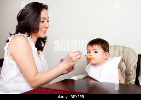 Schöne glückliche Mutter oder Kindermädchen feeds Baby junge Mädchen orange Püree mit Löffel, Baby isst unordentlich, beim Sitzen am Tisch. Stockfoto