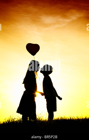 Silhouette der ein junges Mädchen und jungen Händchenhalten und küssen mit einem Herz-Form-Ballon bei Sonnenuntergang. Stockfoto