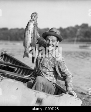 1940S 1950S SIND STOLZ DARAUF, GLÜCKLICHER MANN ANGELN VON EINEM RUDERBOOT LÄCHELND HÄLT NUR MIT GEFANGENEN FISCHE Stockfoto