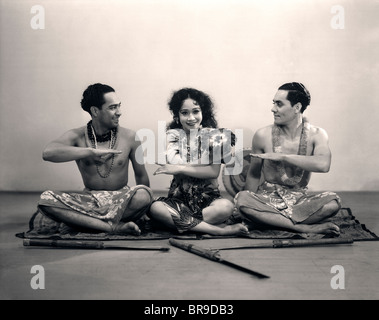 1920ER JAHREN ZWEI MÄNNER SITZEN AUF BODEN TUN, POLYNESISCHE TANZ MIT HANDGESTEN FRAU BLICK IN DIE KAMERA SCHAUEN Stockfoto