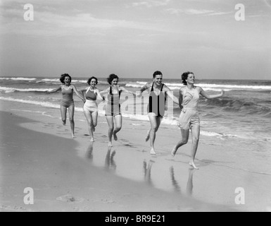 1930ER JAHREN VIER FRAUEN UND EIN MANN AM STRAND HAND IN HAND LAUFEN Stockfoto