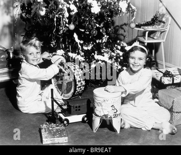 1950ER JAHREN LÄCHELNDEN JUNGEN UND MÄDCHEN MIT SPIELZEUG UNTER BAUM WEIHNACHTSMORGEN Stockfoto