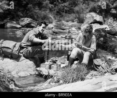 1930ER JAHREN PAAR MIT PICKNICK, SITZEN AUF DEN FELSEN BACH MANN GIEßEN DRINK FÜR FRAU AUS THERMOSKANNE Stockfoto