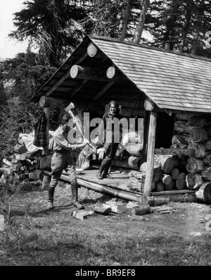 1920S 1930S PAAR FRAU STAND AUF DER VERANDA DER BLOCKHÜTTE MIT KRUG MANN HACKEN HOLZ ASSINIBOINE KANADA Stockfoto