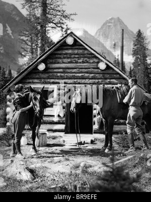 1920S 1930S PAAR VOR BLOCKHAUS BEREITZUSTEHEN PFERDE MIT SÄTTEL TRAGEN REITHOSE ASSINIBOINE KANADA Stockfoto