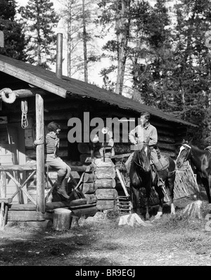 1920S 1930S PAAR VOR BLOCKHÜTTE FRAU SITZT AUF DER VERANDA GELÄNDER MANN AUF PFERD ALBERTA KANADA Stockfoto