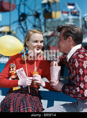 1940S 1950S JUNGES PAAR AM VERGNÜGUNGSPARK MANN ESSEN POPCORN FRAU MIT BALLON UND TRINKEN UND CRACKERJACK BOX Stockfoto