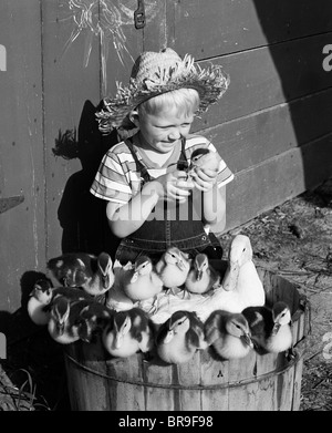 1950ER JAHREN BAUERNJUNGE HOLDING ENTLEIN SITZEN KORB GEFÜLLT MIT ENTENMUTTER UND BABYS Stockfoto