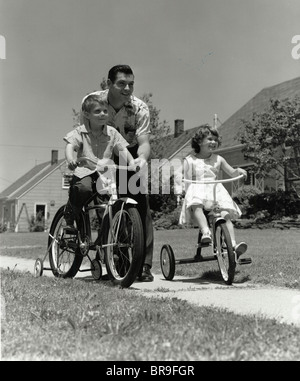 1950ER JAHREN VATER DRÄNGEN JUNGE AUF FAHRRAD MIT STÜTZRÄDERN UND MÄDCHEN AUF DREIRAD NACHBARSCHAFT BÜRGERSTEIG Stockfoto