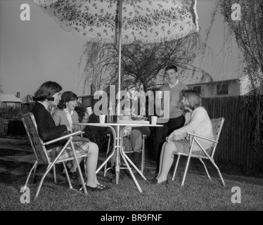 1960ER JAHRE GRUPPE TEENAGER SITZEN IM HINTERHOF UNTER RASEN REGENSCHIRM ESSEN SNACKS TRINKEN LIMONADE Stockfoto