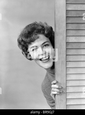 1960ER JAHREN LÄCHELNDE FRAU UM LUFTSCHLITZ HOLZTÜR BLICK IN DIE KAMERA GUCKEND Stockfoto