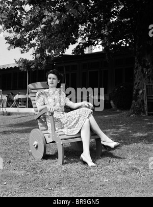 1940ER JAHREN FRAU SITZT IN HÖLZERNEN LIEGESTUHL MIT RÄDERN AUF RASEN VOR HAUS BLICK AB IN FERNE Stockfoto