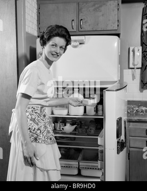 1950ER JAHRE LÄCHELND FRAU HAUSFRAU SETZEN STÜCK BUTTER IN ELEKTRISCHEN KÜHLSCHRANK KÜCHE MIT BLICK IN DIE KAMERA Stockfoto