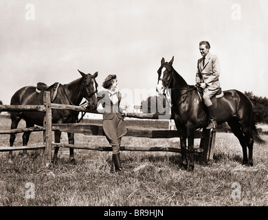 1930S 1940S PAAR IN RIDING GEAR MANN REITEN PFERD FRAU STAND VON HOLZZAUN Stockfoto