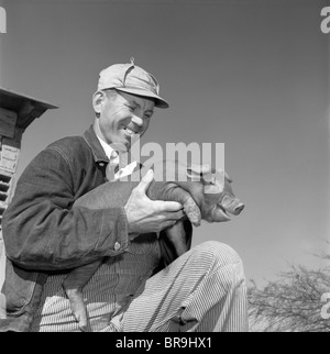 1950ER JAHREN LÄCHELND MANN BAUER TRAGEN HUT ARBEIT JACKE DENIM GESTREIFT OVERALLS HALTEN DUROC FERKEL AUF DIE KNIE Stockfoto