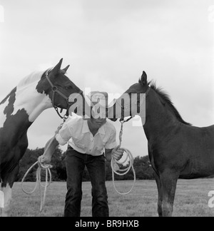 1960ER JAHRE JUNGE COWBOY HUT HÄLT SEILE ZÜGEL HARNESS HALTER VON 2 PFERDEN EIN AUF BEIDEN SEITEN DES GESICHTES KÜSSEN IHN LUSTIG Stockfoto