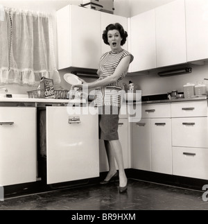1950ER JAHREN FRAU HAUSFRAU TRAGEN KITTEL SETZEN GESCHIRR IN SPÜLMASCHINE IN EINEM VORORT KÜCHE, BLICK IN DIE KAMERA Stockfoto