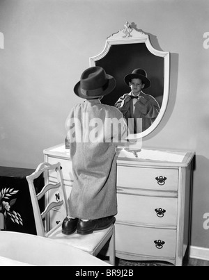 1950ER JAHRE JUNGE GEKLEIDET IN DES VATERS HUT MANTEL & SCHUHE AUF STUHL IN SPIEGEL Stockfoto