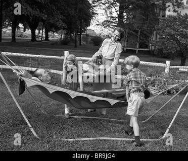 1950ER JAHREN MUTTER & KINDER SERVIEREN VATER IN HÄNGEMATTE Stockfoto