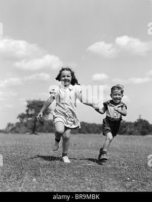 1940ER JAHREN ZWEI LÄCHELNDE KINDER LAUFEN HAND IN HAND IN WIESE Stockfoto