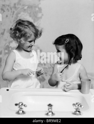 1930ER JAHREN ZWEI MÄDCHEN IM BADEZIMMER WASCHBECKEN HALTEN ZAHNBÜRSTE Stockfoto