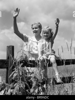 1930ER JAHREN MÄDCHEN SITZEN AUF ZAUN MIT FRAU NEBEN IHR IM FELD WINKEN Stockfoto