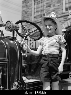 1930ER JAHREN LÄCHELNDE JUNGE STAND NEBEN FEUERWEHRAUTO TRAGEN FIREMANS HUT Stockfoto