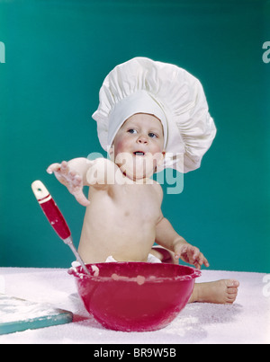 1960ER JAHRE BABY TRAGEN KOCH HUT MIT ROTER MISCHENDEN SCHÜSSEL UND LÖFFEL LUSTIGE MIMIK Stockfoto