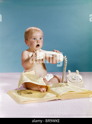 1960ER JAHRE BABY ÜBERRASCHT GESICHTSAUSDRUCK HÄLT HAND TELEFONAPPARAT ÜBER TELEFONBUCH BRANCHENBUCH Stockfoto