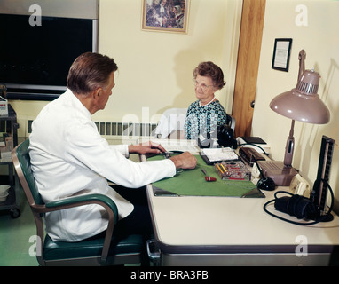 1960ER JAHRE ARZTPRAXIS MIT ARZT IM GESPRÄCH MIT SENIOR WOMAN PATIENT Stockfoto
