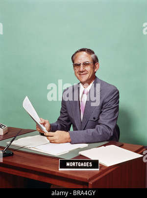 1970ER JAHREN LÄCHELND MANN AM SCHREIBTISCH MIT HYPOTHEK OFFIZIER ZEICHEN HOLDING PAPIERE Stockfoto