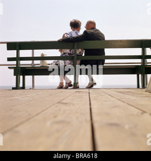 1970 1970S MANS RETRO-RÜCKANSICHT ÄLTERES PAAR SITZT AUF DER BANK MIT BLICK AUF WASSER ARM UM WOMANS SCHULTERN Stockfoto