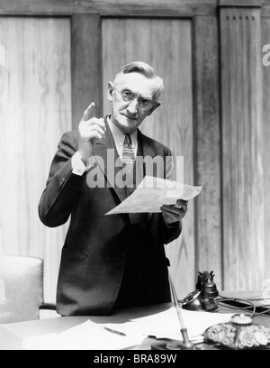 1930ER JAHRE ÄLTERER MANN IM BÜRO STEHEN HINTER SCHREIBTISCH GESTIKULIEREN MIT ANGEHOBENEN FINGER Stockfoto