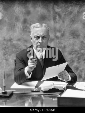 1930ER JAHRE ÄLTEREN MANN SITZEN AM SCHREIBTISCH IM BÜRO ZEIGEN MIT STRENGEN AUSDRUCK Stockfoto