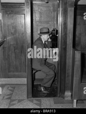 1940ER JAHRE MANN SITZT IN HÖLZERNEN TELEFONZELLE IN EMPFÄNGER SPRECHEN Stockfoto