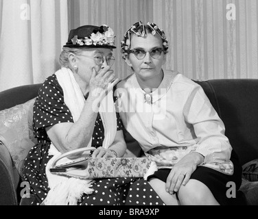 1960ER JAHRE ÄLTERE FRAUEN AUF COUCH AUSTAUSCH VON GEHEIMEN EIN IN GEBLÜHT HAT MAN IN LOCKENWICKLER Stockfoto