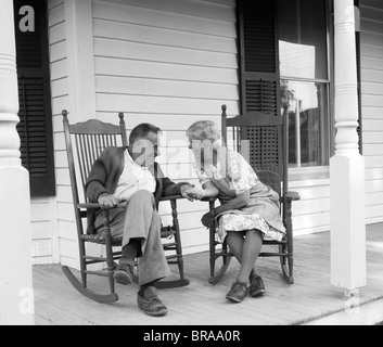 1970ER JAHRE ÄLTERES EHEPAAR IN ROCKING STÜHLE AUF DER VERANDA HAND IN HAND Stockfoto