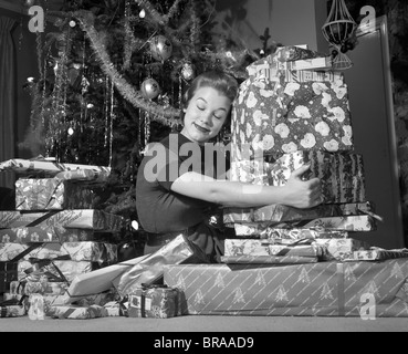 1950ER JAHREN FRAU SITZT VON WEIHNACHTSBAUM UMARMT HAUFEN VON GESCHENKEN Stockfoto