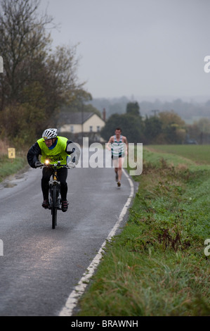 Eine offizielle Rennen auf einem Fahrrad Schlachten Wind und Regen, wie er die einsame Spitze Läufer Billericay 10 km-Straßenlauf begleitet. Stockfoto