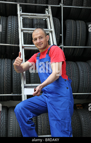Ein Lächeln auf den Lippen Mechaniker in einer Garage stehen einem eine Leiter neben einem Rack voller Reifen und eine positive Geste. Stockfoto