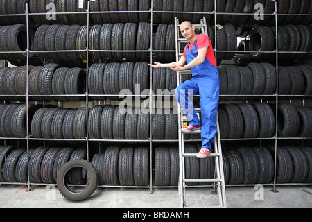 Ein Lächeln auf den Lippen Mechaniker in einer Garage stehen einem eine Leiter neben einem Rack voller Reifen und eine positive Geste. Stockfoto