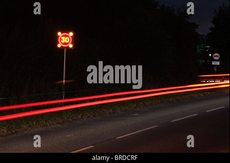 30 km/h Geschwindigkeit Zeichen blinkt in ein Dorf und Auto Rückleuchten in der Nacht. Langzeitbelichtung Stockfoto